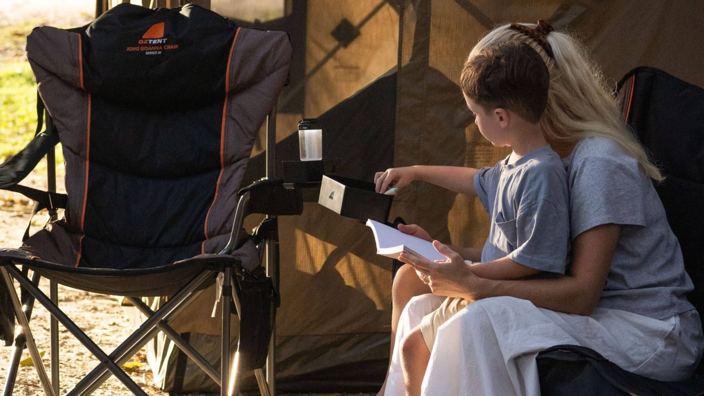 Child seated beside an adult in a camping setup, writing in a notebook with a tent backdrop nearby.